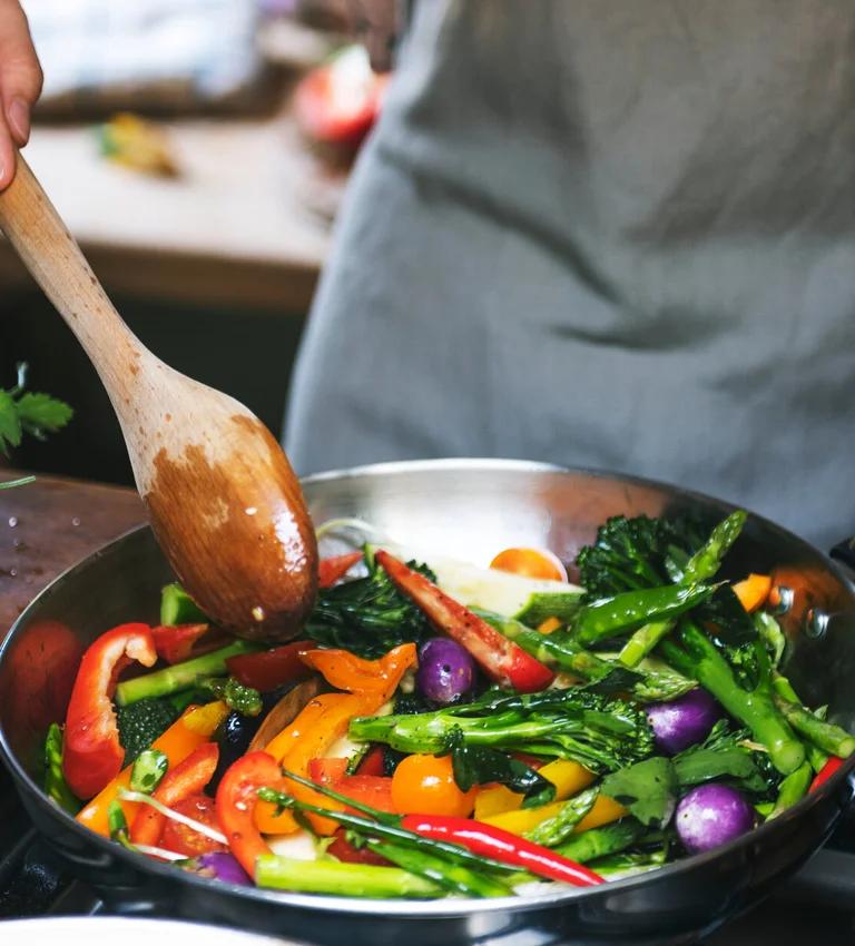 Person brät Gemüse in einer Pfanne und rührt mit einem Holzlöffel.