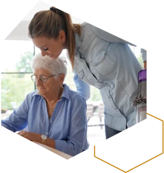 Pflegekraft unterstützt eine ältere Frau beim Ausfüllen von Unterlagen am Tisch.