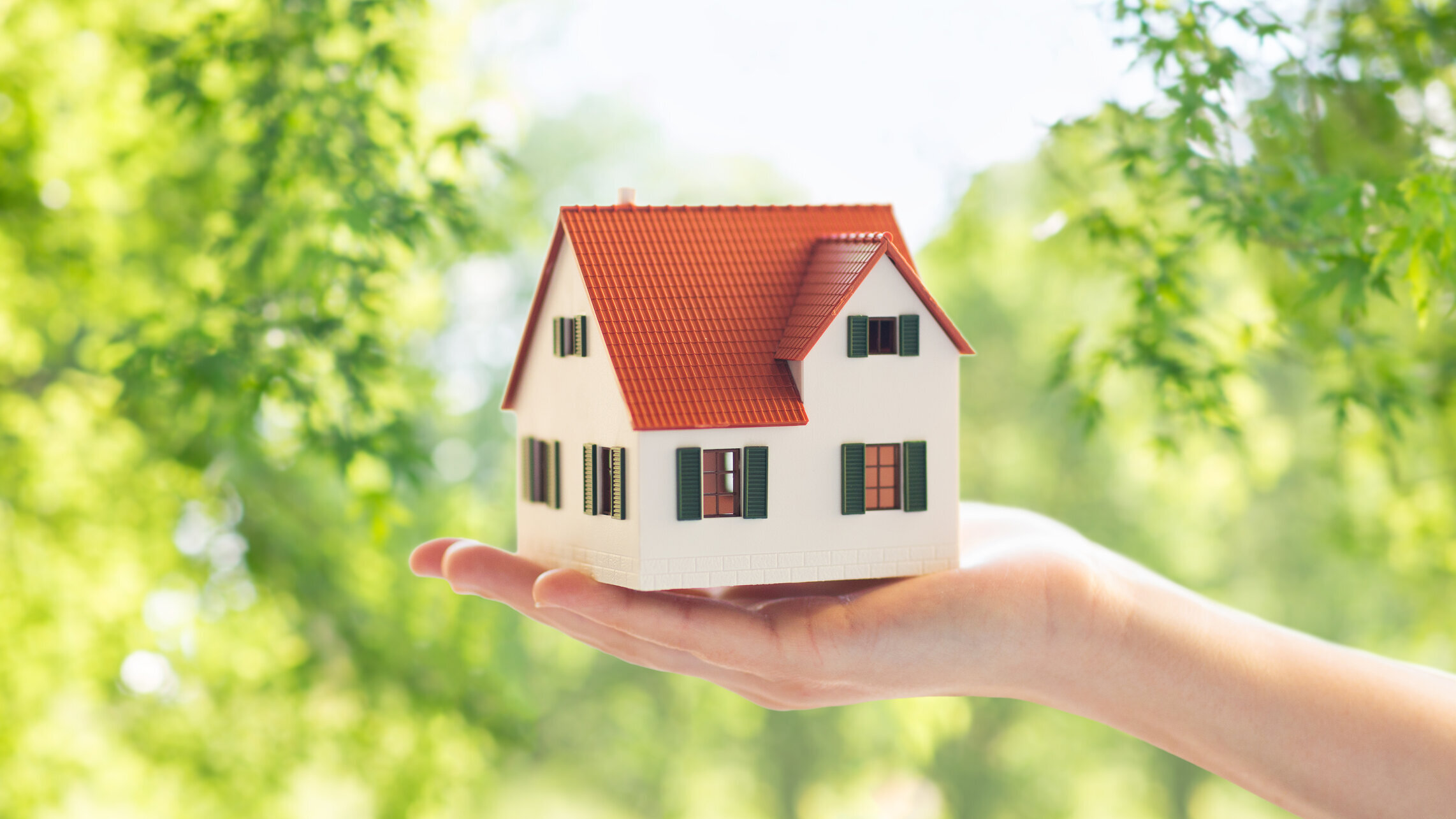 „Hand hält ein kleines Hausmodell vor grünem Hintergrund.