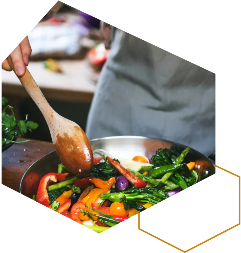 Person brät Gemüse in einer Pfanne und rührt mit einem Holzlöffel.“